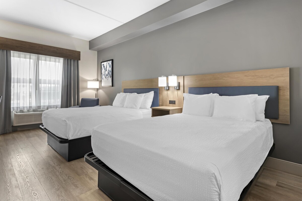 A hotel room with two beds, a large window with curtains, a wall-mounted artwork, and a bedside table with a lamp.