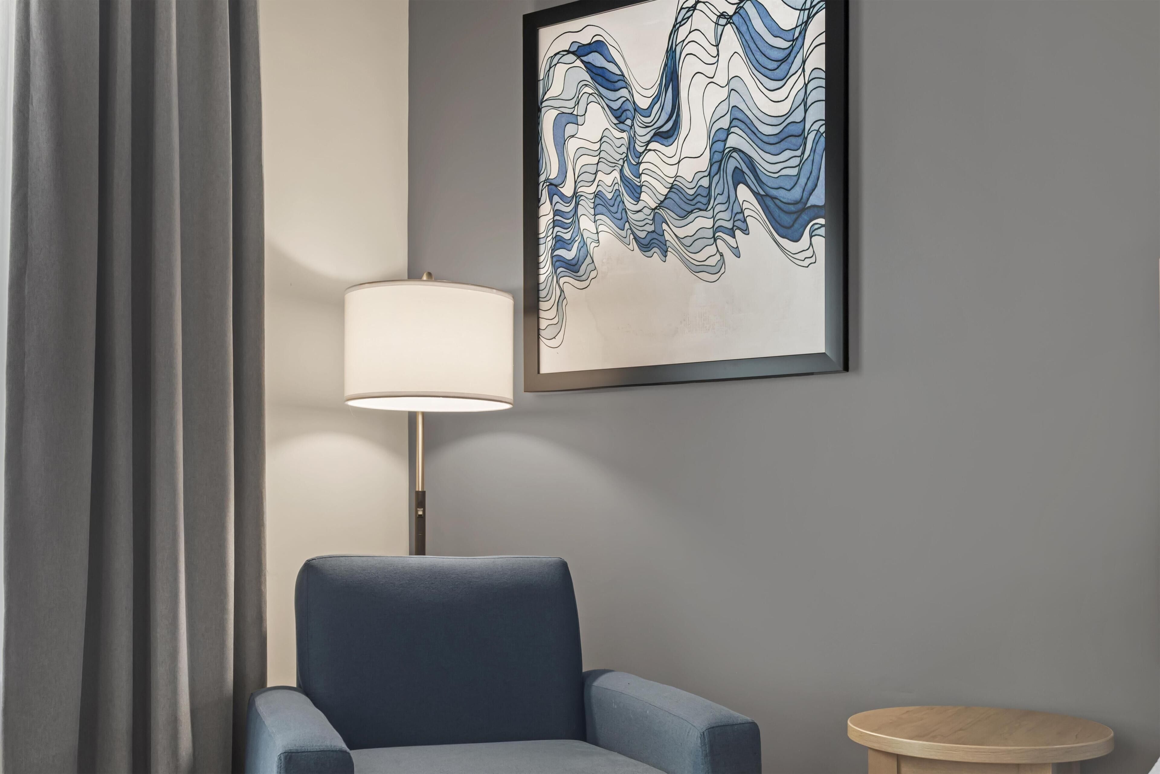 Une pièce avec un fauteuil bleu, un lampadaire, une petite table ronde et un tableau abstrait encadré accroché au mur.