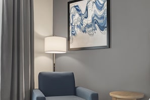 Una habitación con un sillón azul, una lámpara de pie, una mesita redonda y un cuadro abstracto enmarcado en la pared.