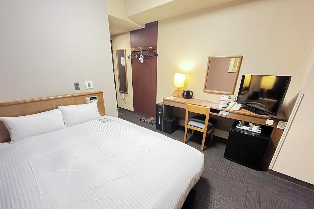 A hotel room with a large bed, a desk with a TV, a chair, and a small trash bin.