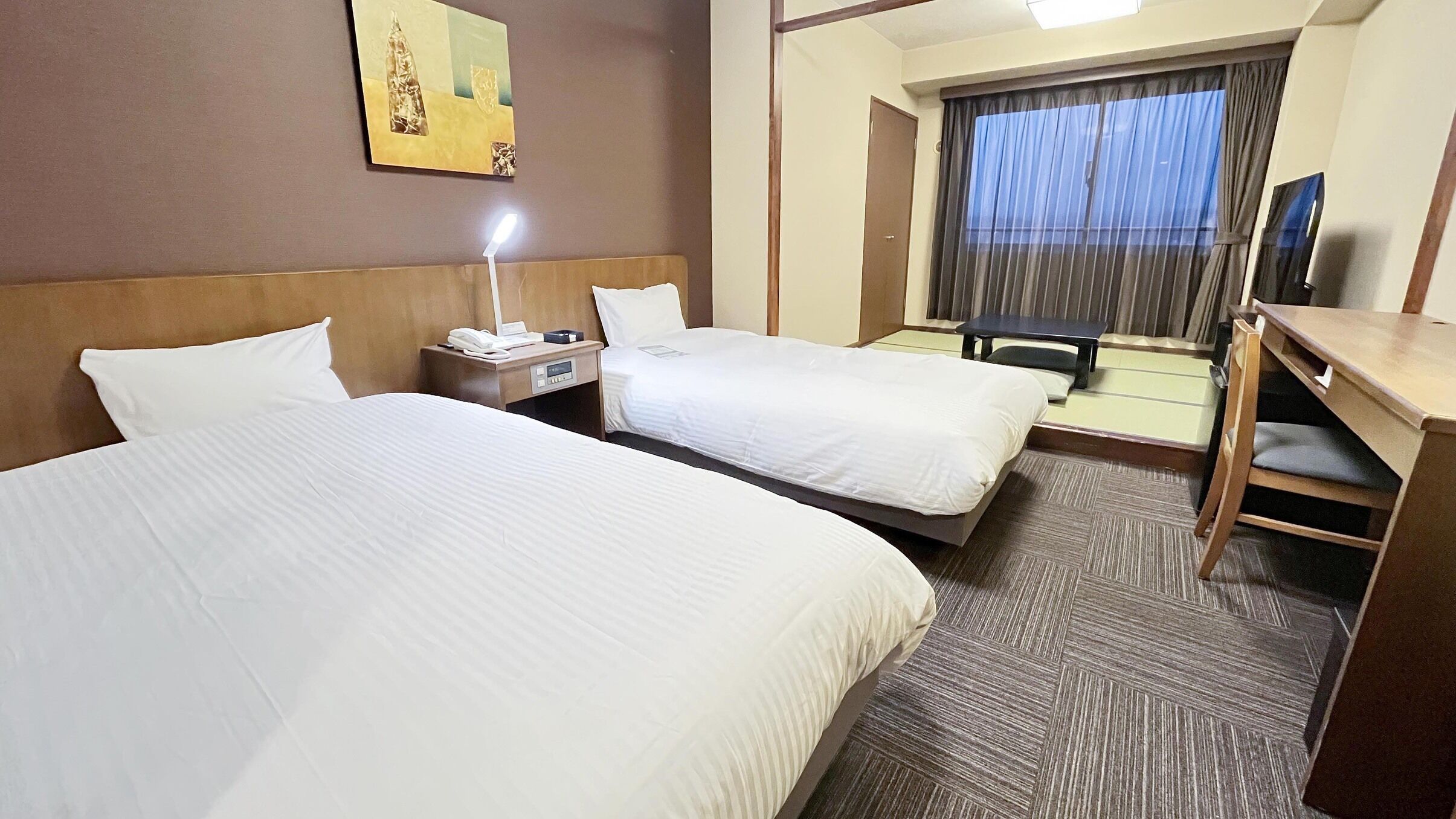 Ein Hotelzimmer mit zwei Betten, einem Schreibtisch, einem Stuhl, einem Fernseher und einem Fenster mit Vorhängen.