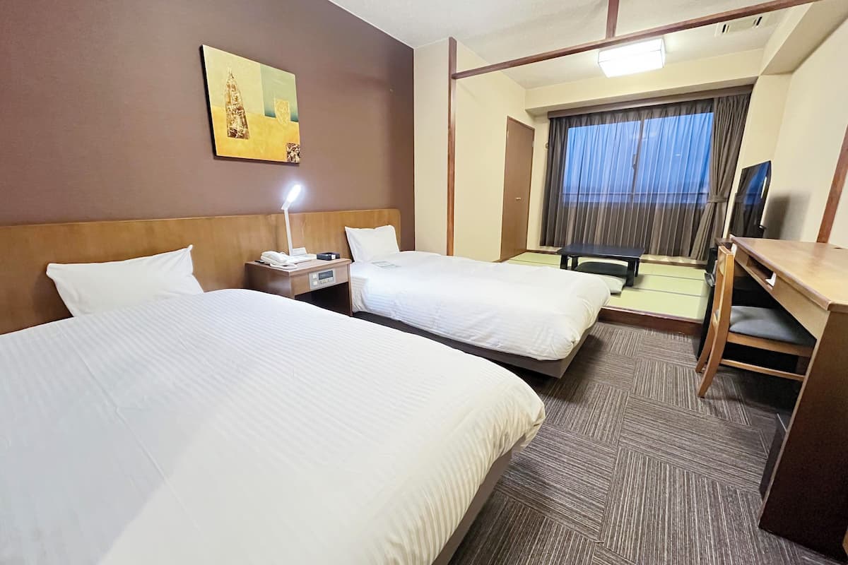 A hotel room with two beds, a desk, a chair, a nightstand with a lamp, a painting on the wall, and a window with curtains.
