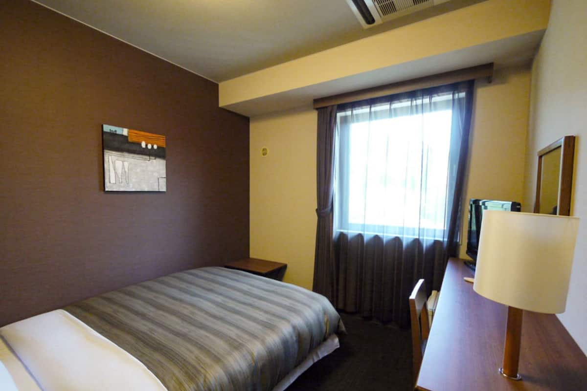 A hotel room with a bed, a desk, a chair, a window with curtains, a wall-mounted picture, and a lamp.