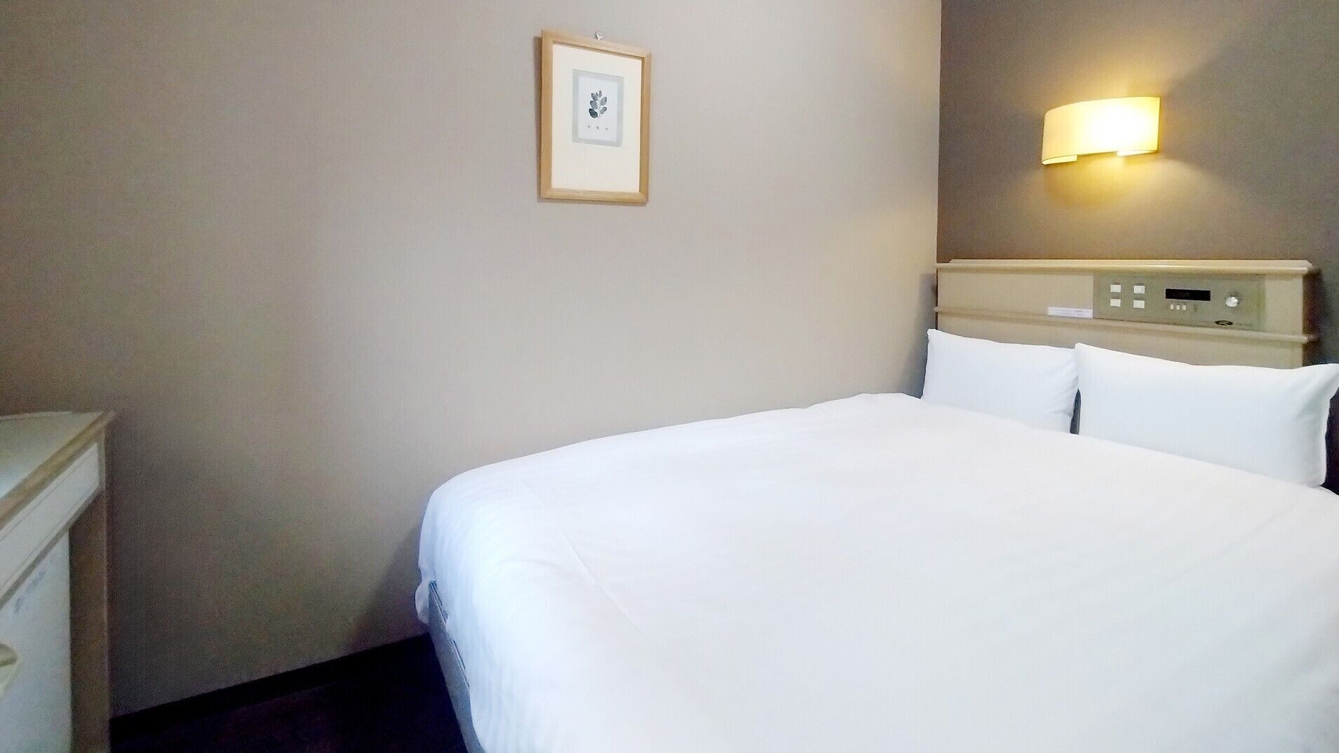 A hotel room with a bed, a bedside table, a wall-mounted light, and a framed picture on the wall.