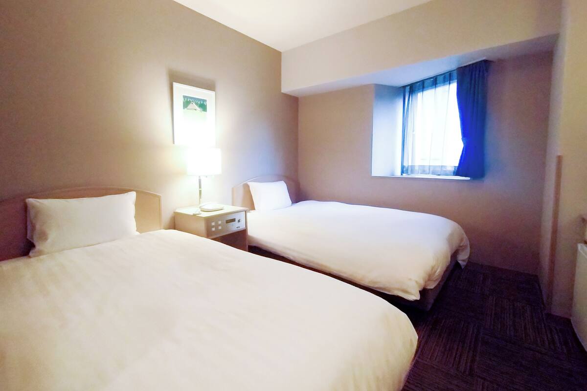 Une chambre d’hôtel avec deux lits, une table de chevet avec une lampe, une fenêtre avec des rideaux et un tableau accroché au mur.