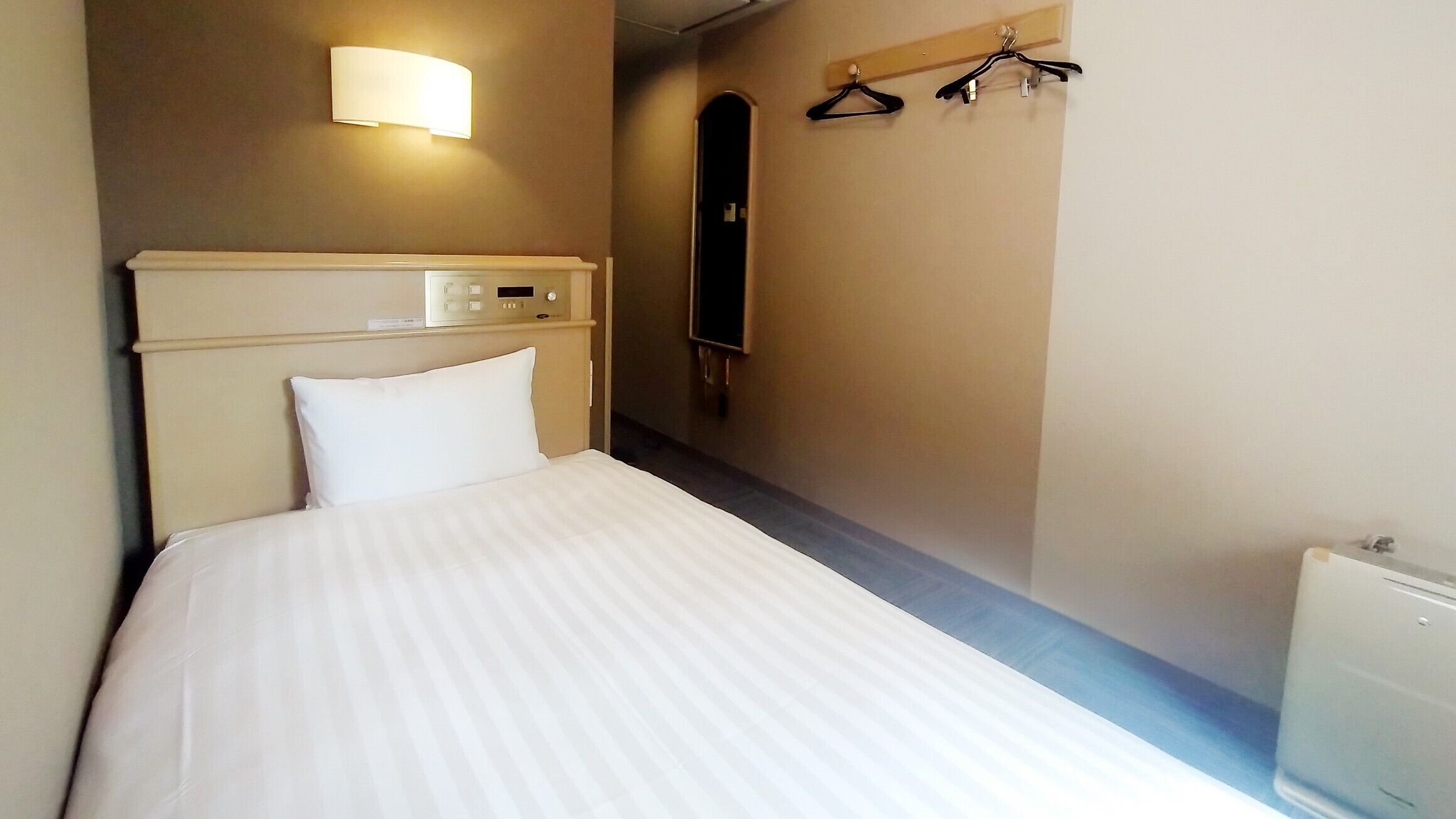 A hotel room with a bed, a control panel, a wall-mounted light, a coat rack, and a mirror.