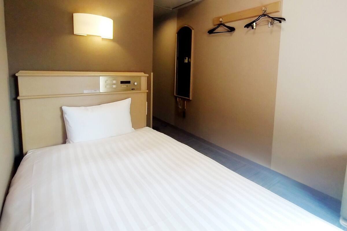 Une chambre d’hôtel comprenant un lit, un panneau de commande, une lampe fixée au mur, un porte-manteau et un miroir.