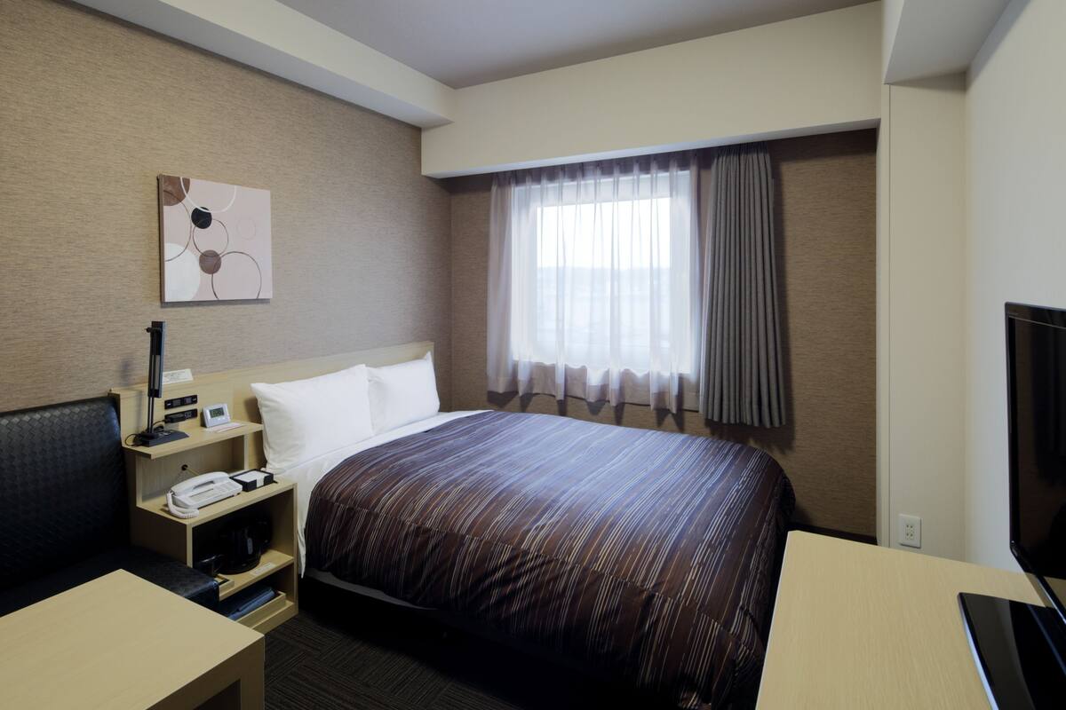 A hotel room with a bed, a desk, a chair, a TV, and a window with curtains.