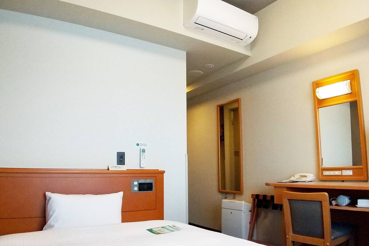 A hotel room with a bed, a desk, a mirror, a phone, and an air conditioning unit.