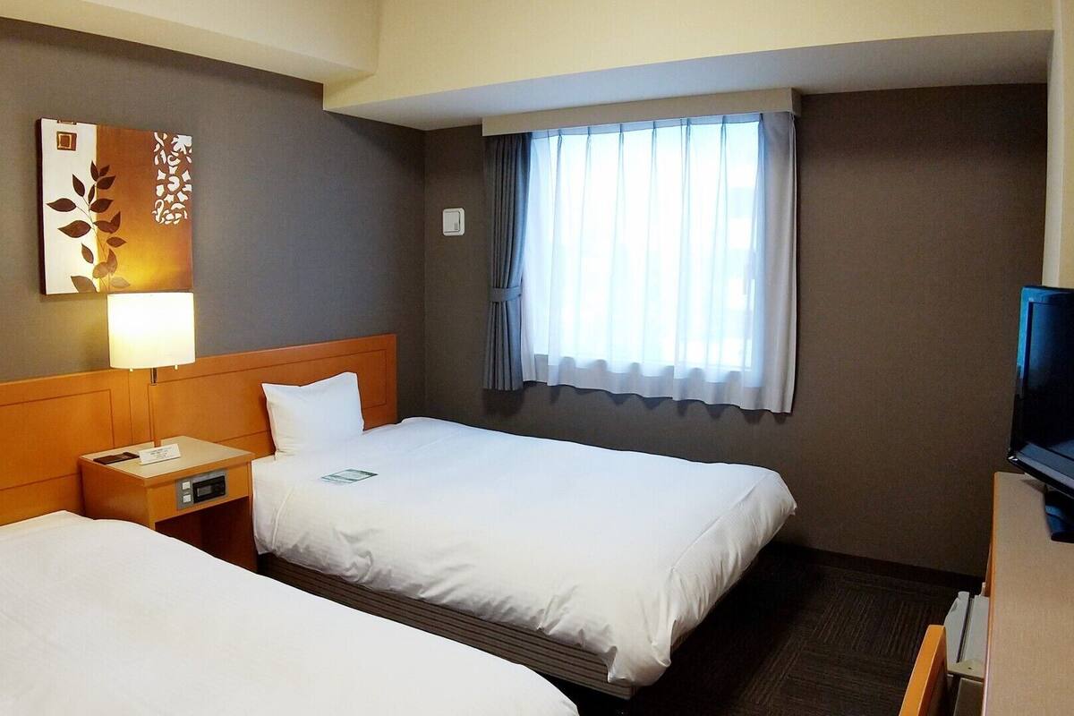A hotel room with two beds, a TV, a lamp, and a window with curtains.