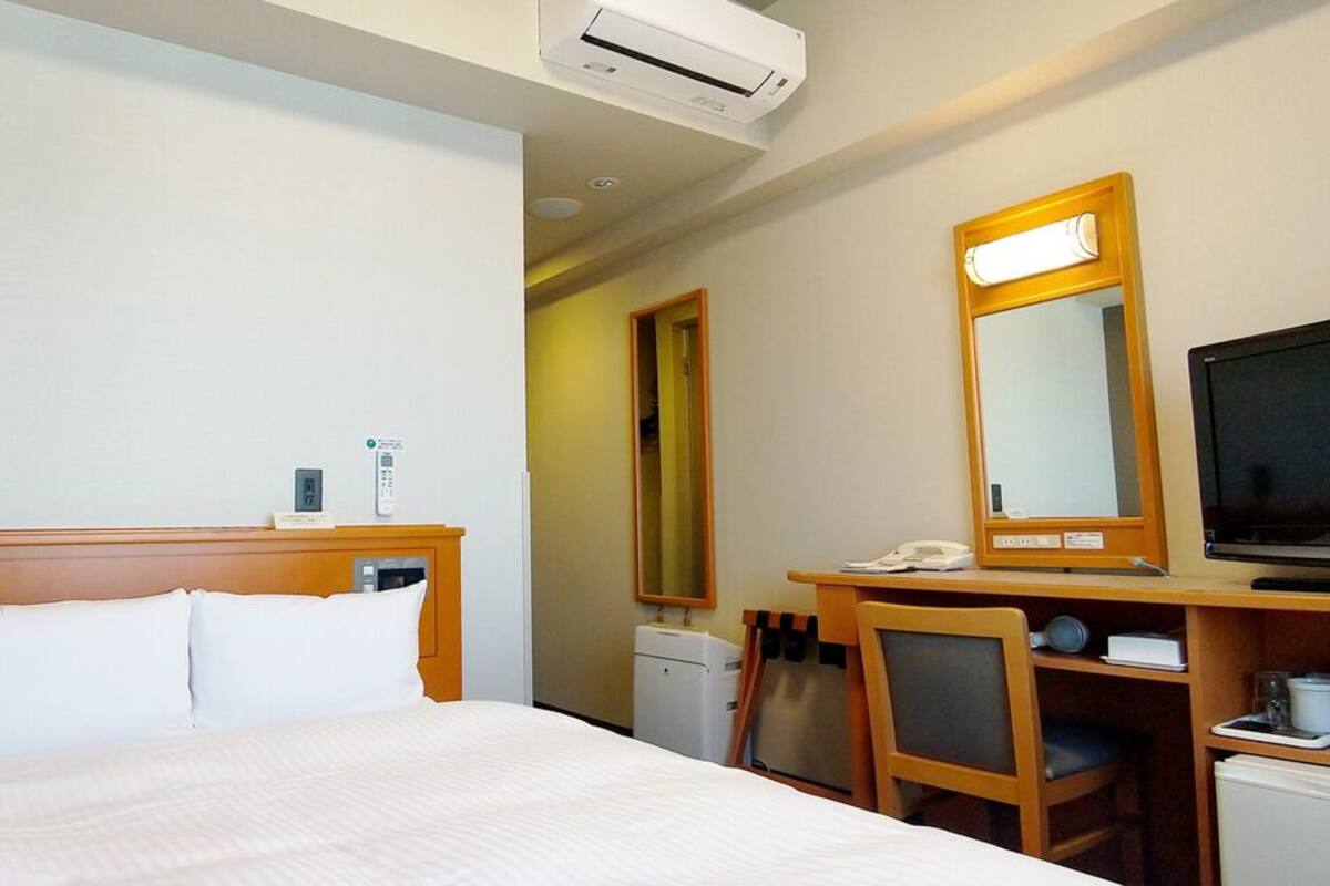 A hotel room with a bed, a desk with a chair, a television, a mirror, and a small kitchenette.