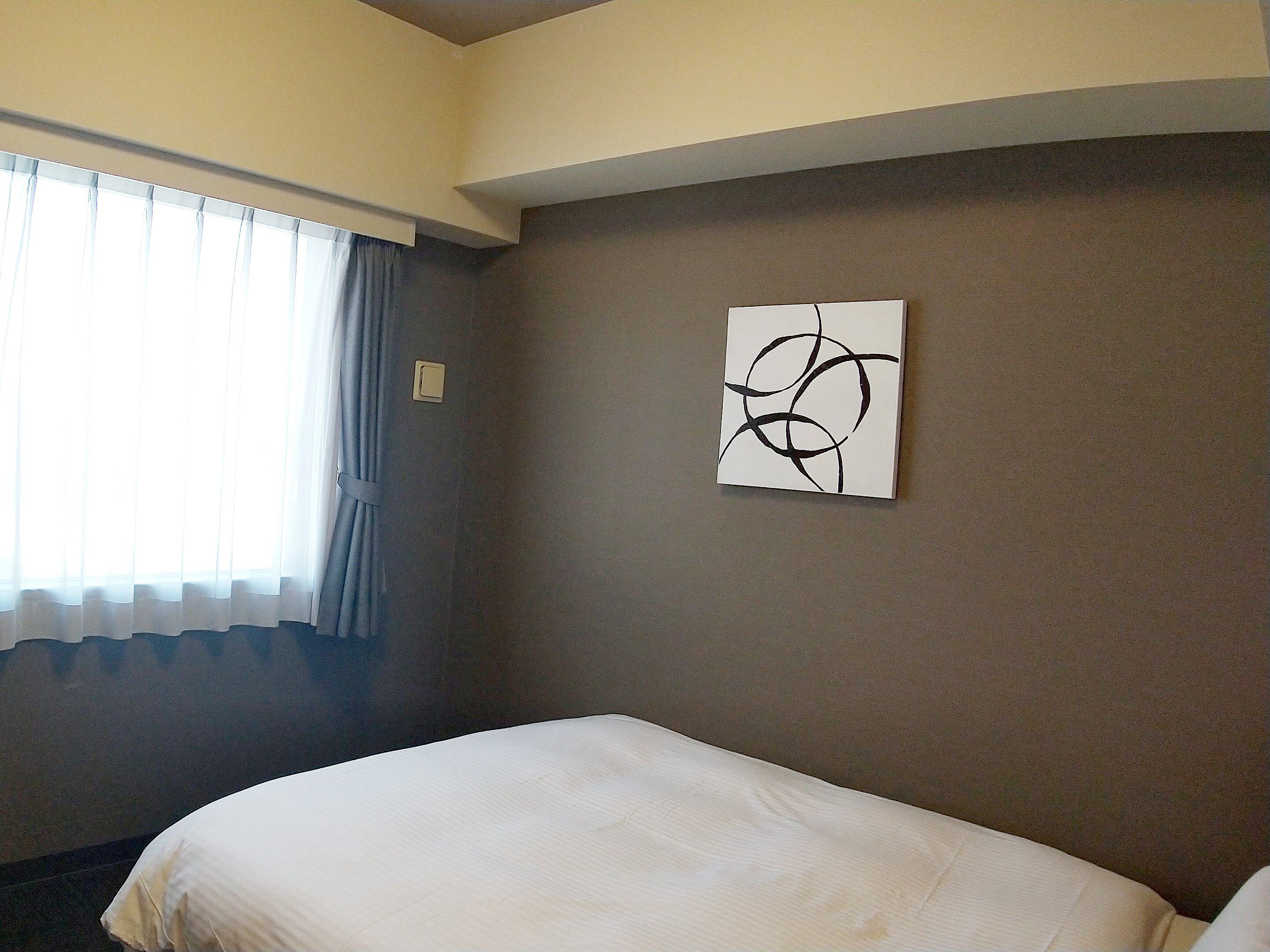 A hotel room with a bed, a window with curtains, and a wall with a piece of abstract art.