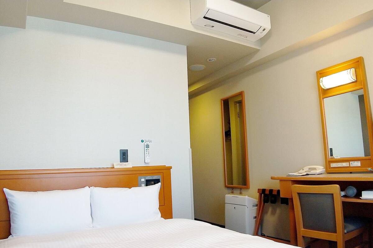 A hotel room with a bed, a desk, a chair, a mirror, a phone, and an air conditioner.