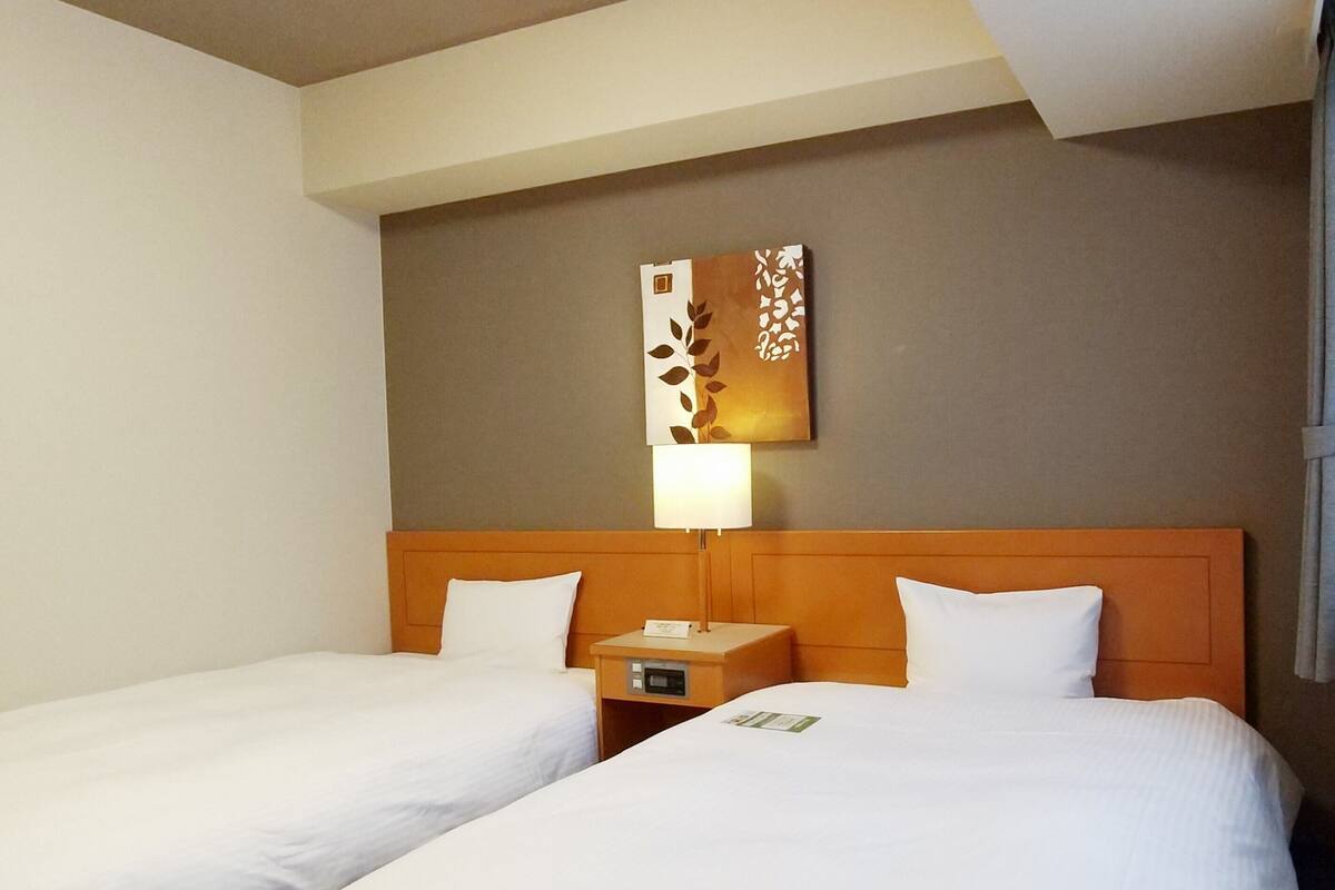 A hotel room with two single beds, a wooden headboard, a nightstand with a lamp, and a wall-mounted artwork.