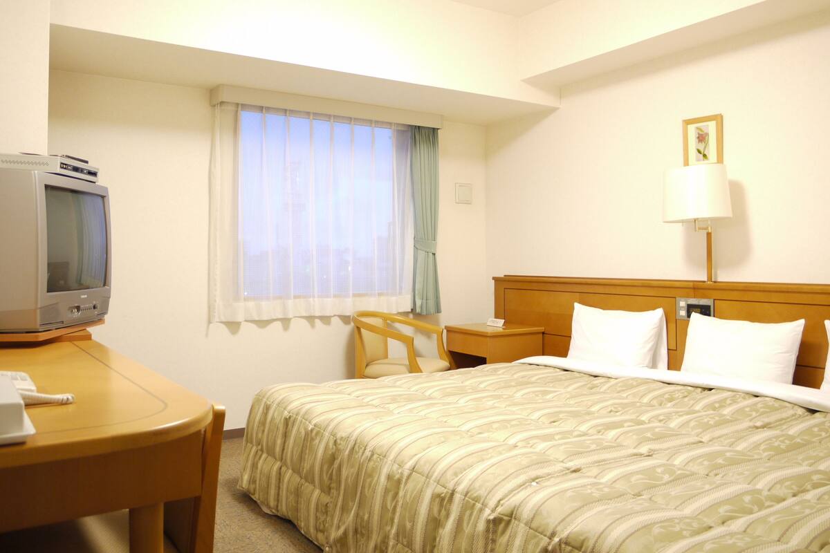 A hotel room with a bed, a desk with a television, a chair, a nightstand, and a lamp.