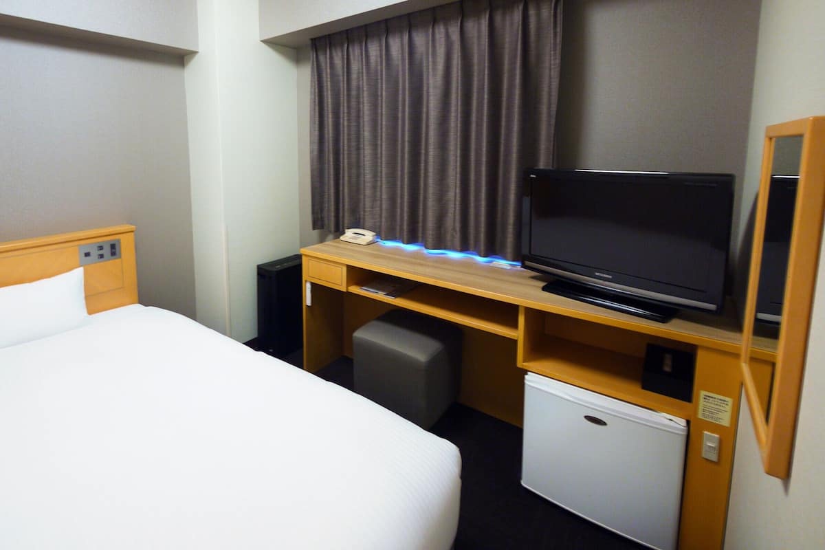 A hotel room with a bed, a desk with a television, a mini-fridge, and a telephone.