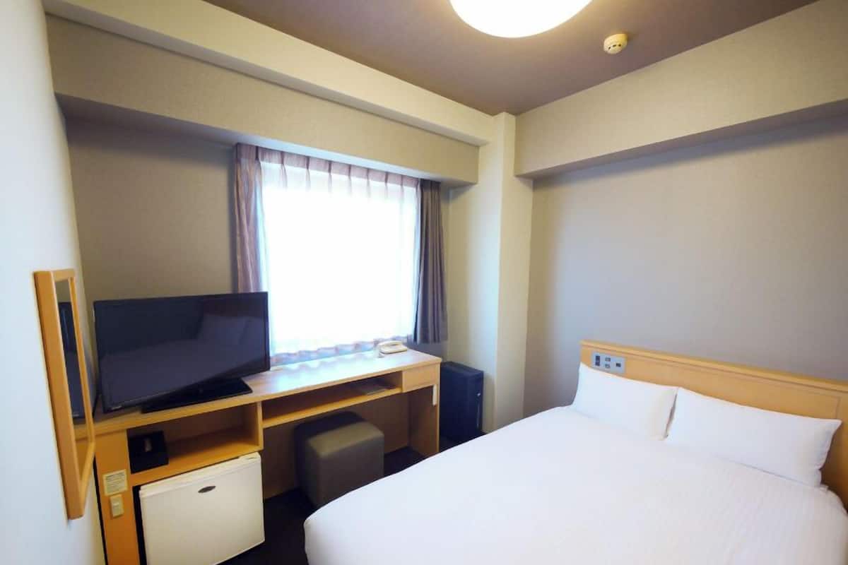A hotel room with a bed, a desk with a television, a small refrigerator, and a window with curtains.