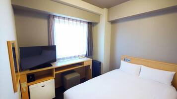 A hotel room with a bed, a desk with a television, a small refrigerator, and a window with curtains.