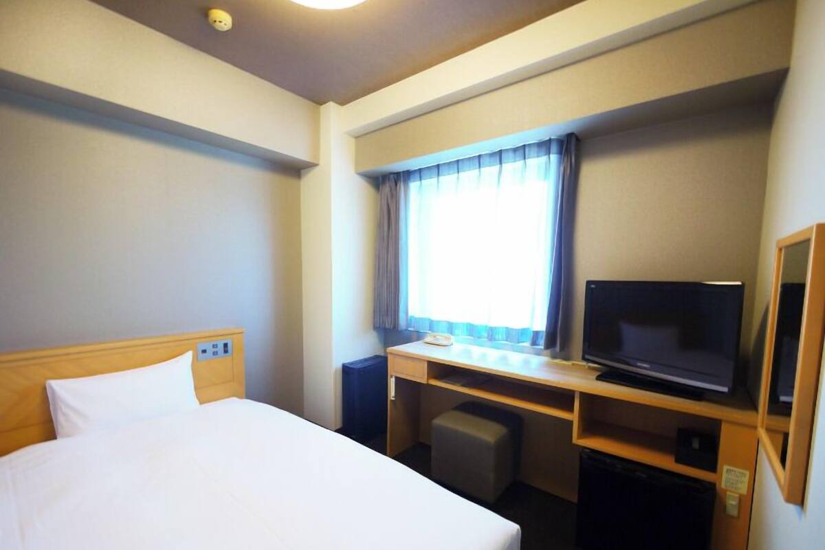 A hotel room with a bed, a desk with a television, a small chair, and a window with curtains.