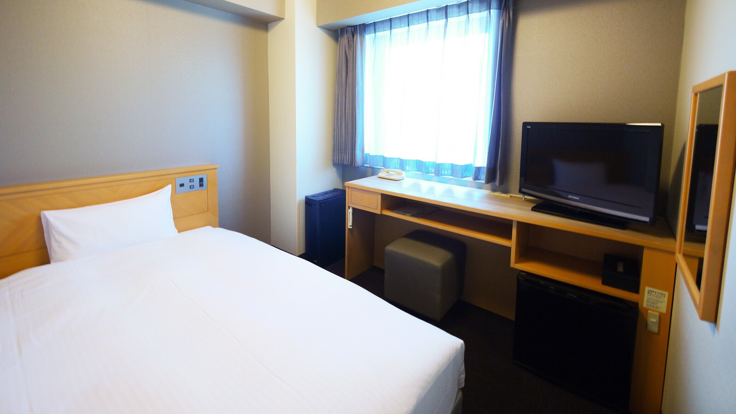 Una habitación de hotel con una cama, un escritorio con televisor, una silla y un pequeño cubo de basura.