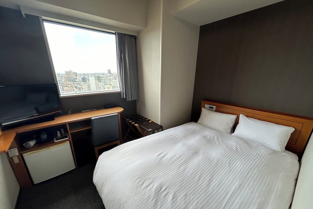 A hotel room with a bed, a desk with a television, a small table, and a window with a view of the city.