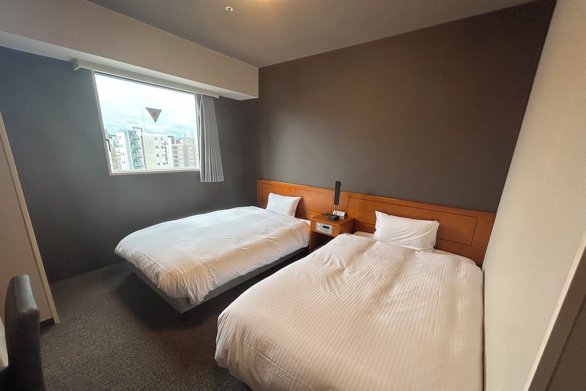 A hotel room with two beds, a nightstand with a lamp, and a window with a view of buildings.