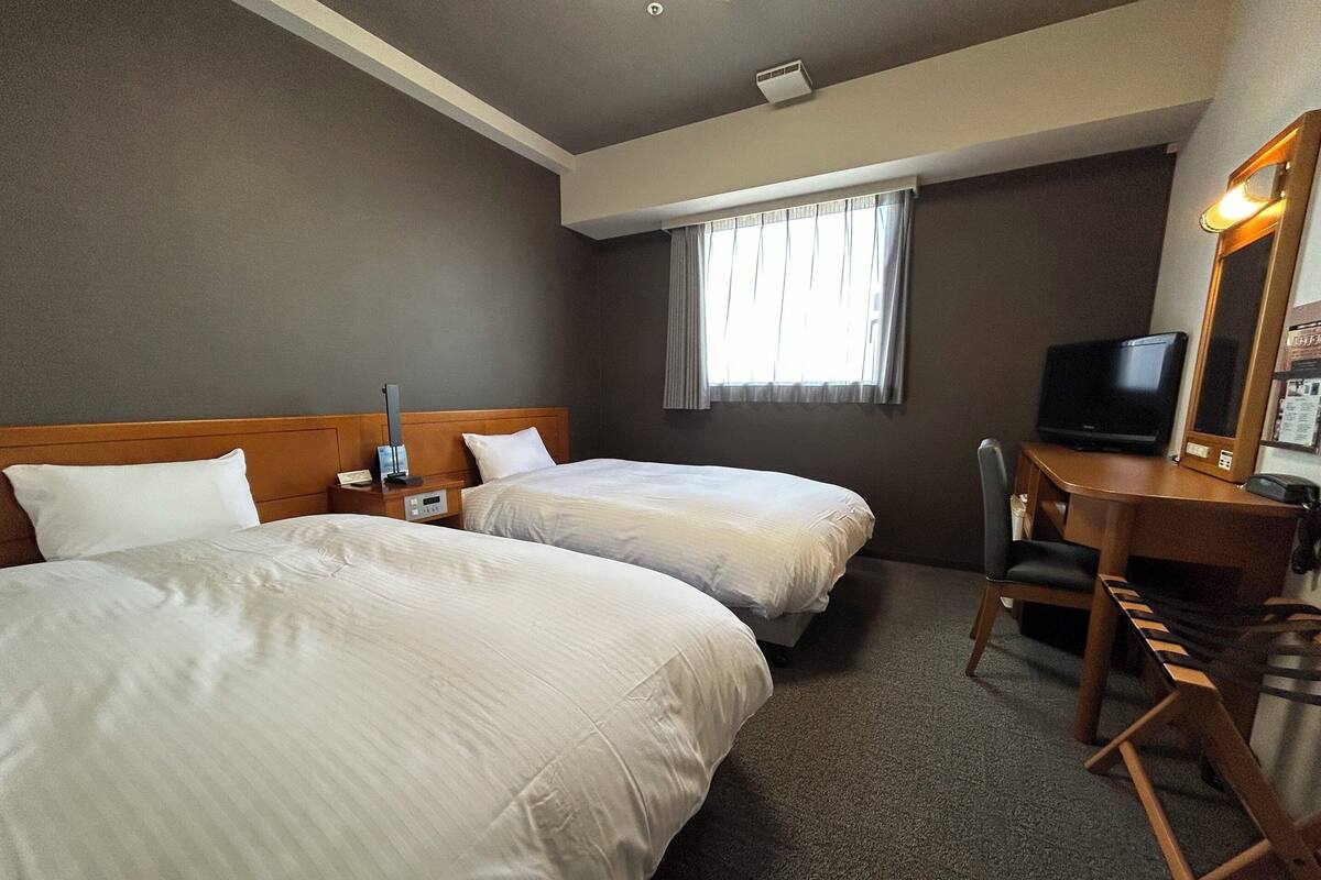 A hotel room with two beds, a desk with a chair, a television, and a window with curtains.