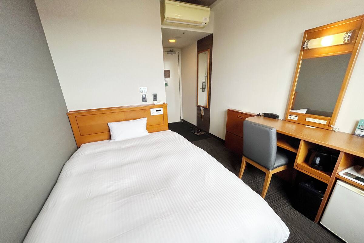 A hotel room with a bed, a desk, a chair, a mirror, and a small cabinet.