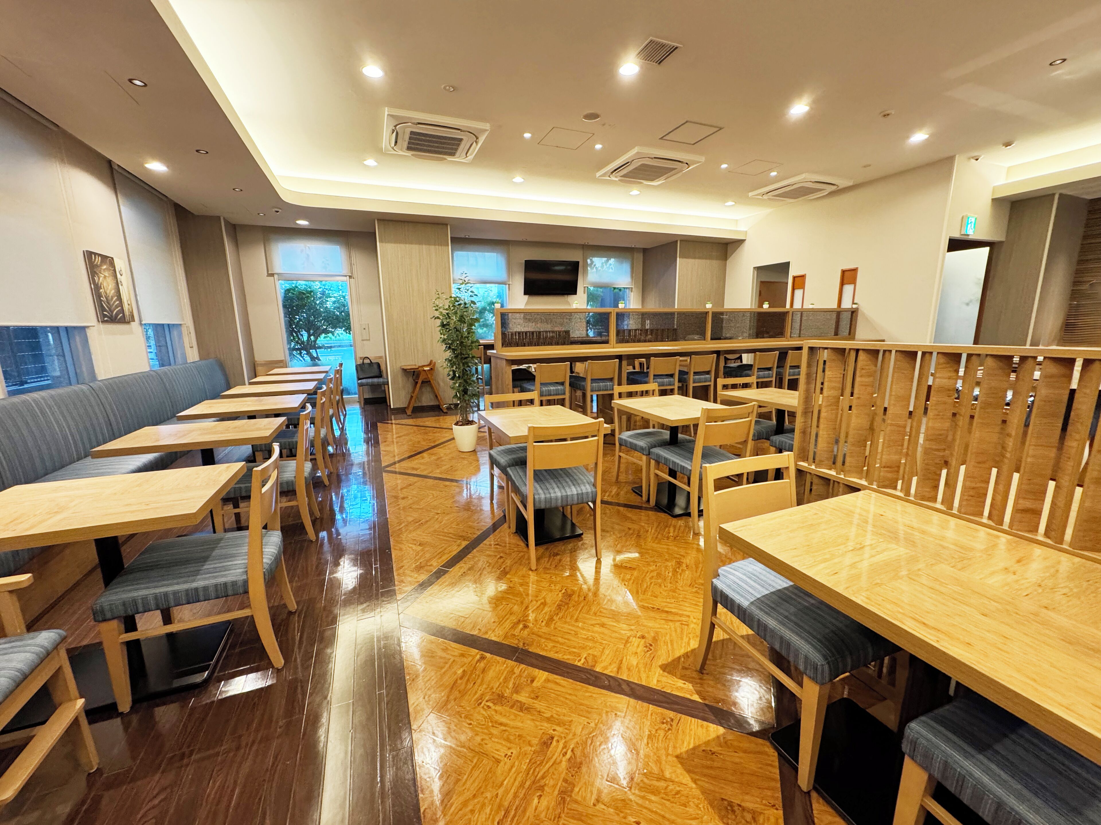 Un restaurante moderno con mesas y sillas de madera, una planta en el centro y una cocina visible a través de una separación.