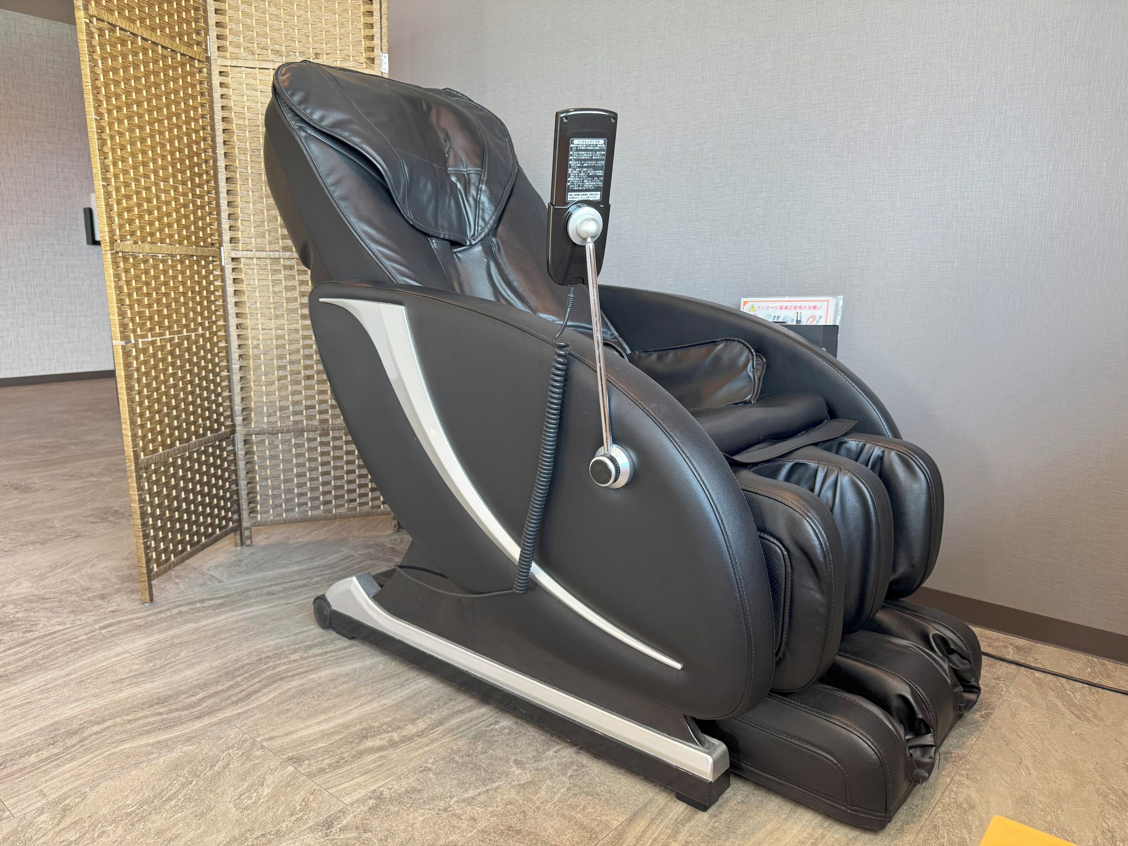 A massage chair with a control panel, positioned in a room with a tiled floor and a woven partition.
