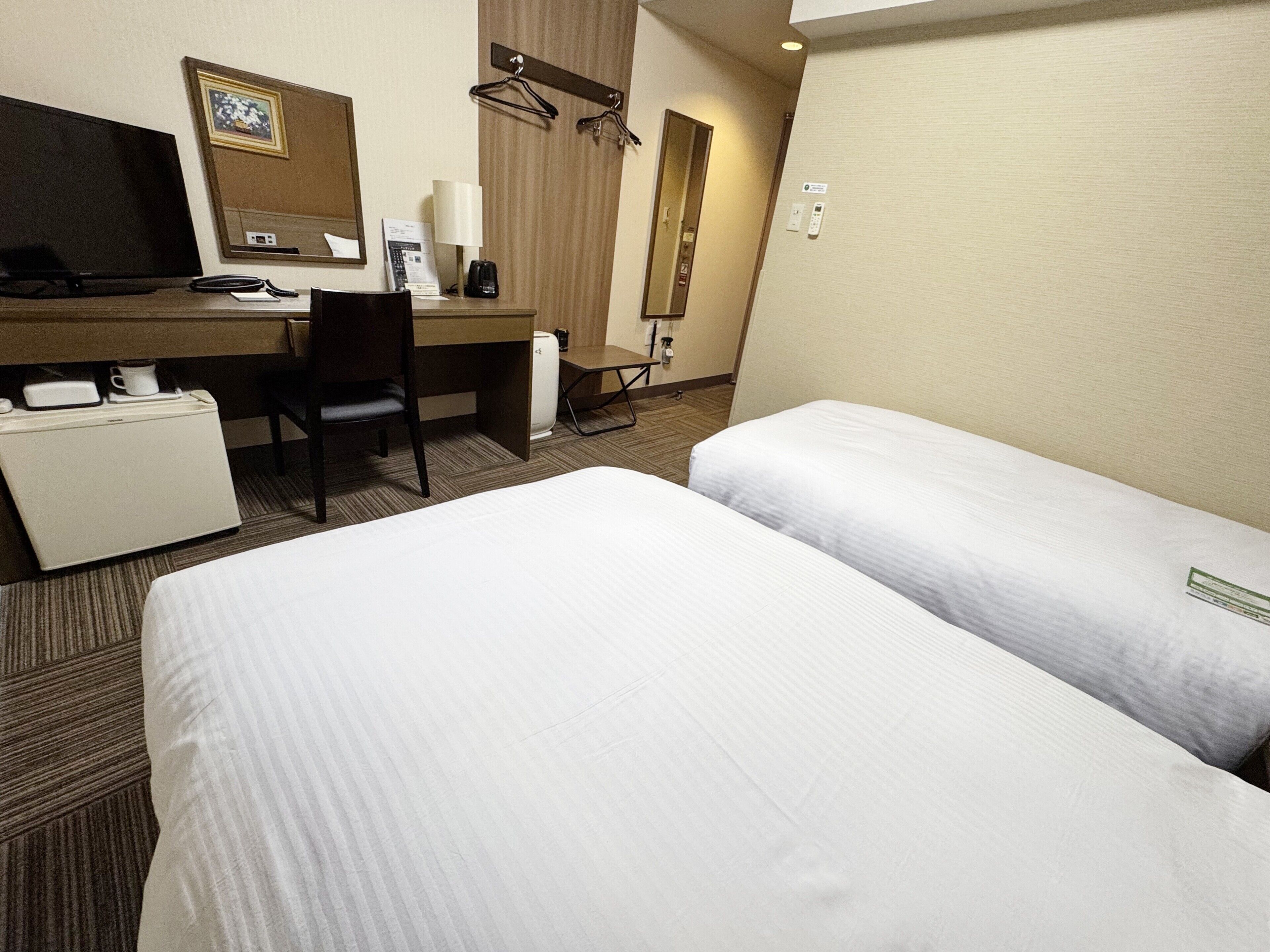 A hotel room with two single beds, a desk with a computer, a chair, a coffee maker, and a small refrigerator.