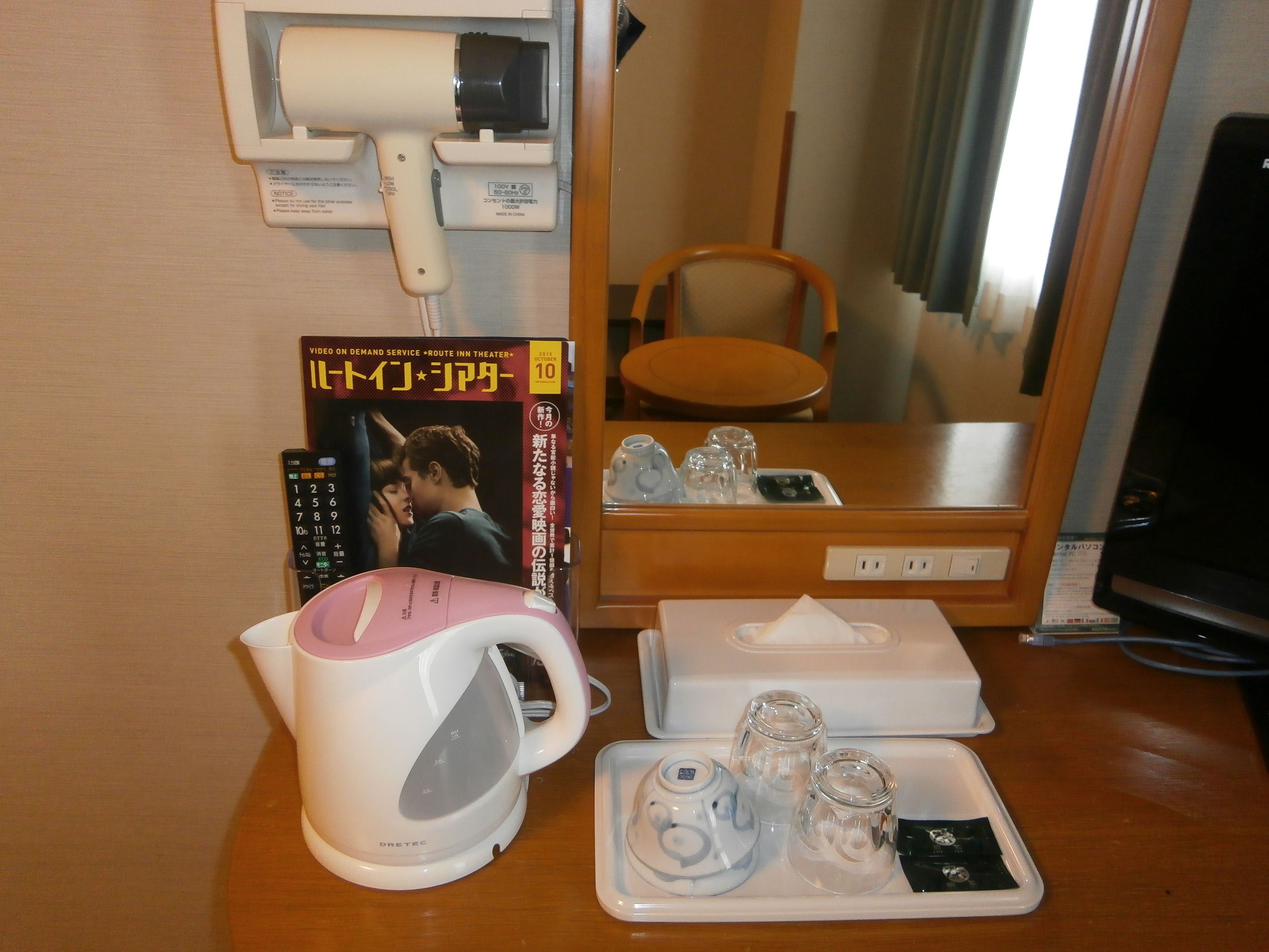 A hotel room with a minibar, a kettle, a tray with glasses, a remote control, and a magazine on the table.