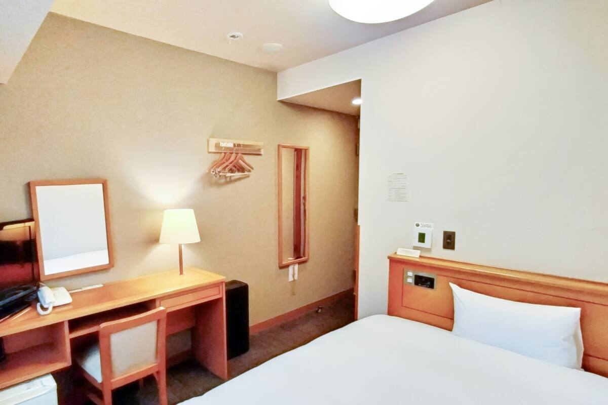 Ein Hotelzimmer mit einem Bett, einem Schreibtisch mit Lampe, einem Spiegel, einem Kleiderhaken und einem kleinen Schrank.