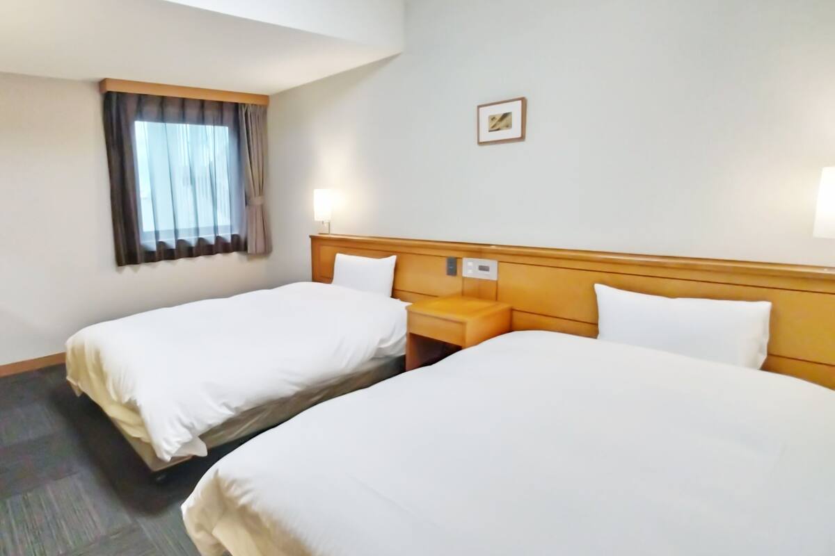 Ein Hotelzimmer mit zwei Einzelbetten, einem Nachttisch, einer Lampe, einem Fenster mit Vorhängen und einem kleinen Bild an der Wand.