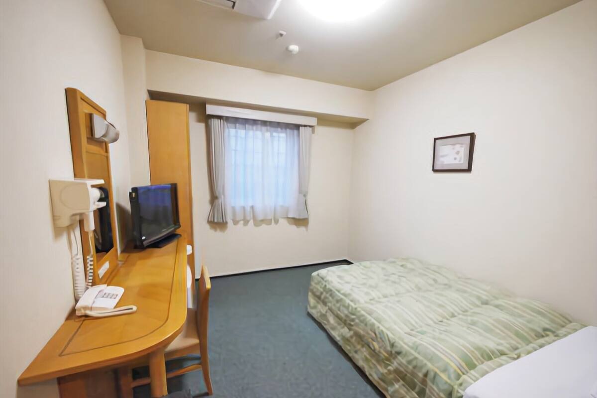 A hotel room with a desk, chair, television, bed, window with curtains, and a framed picture on the wall.