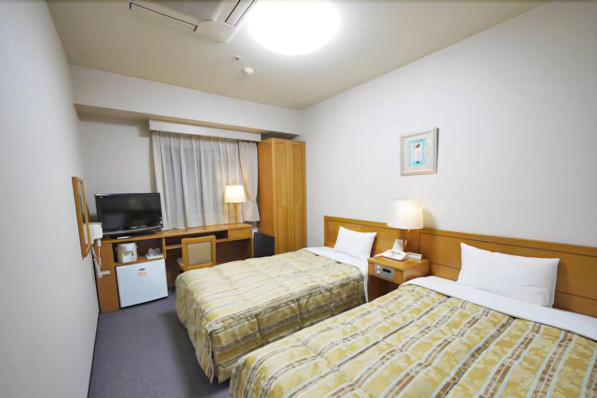 A hotel room with two beds, a desk, a television, and a window with curtains.