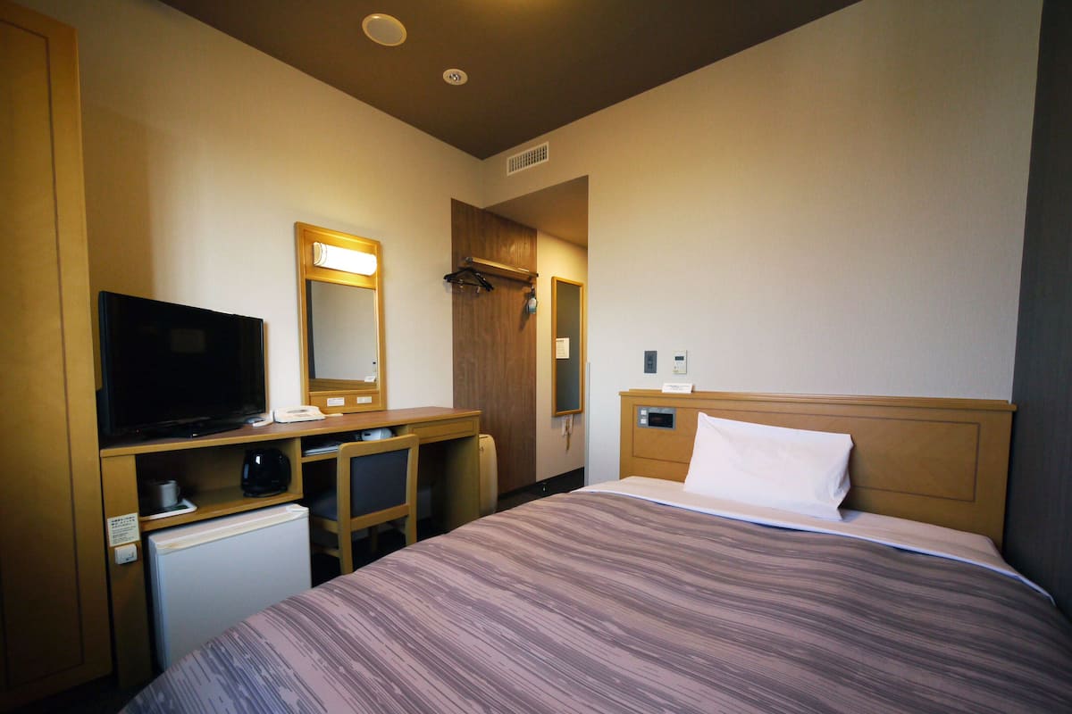 A hotel room with a bed, a desk with a television, a mirror, and a wardrobe.