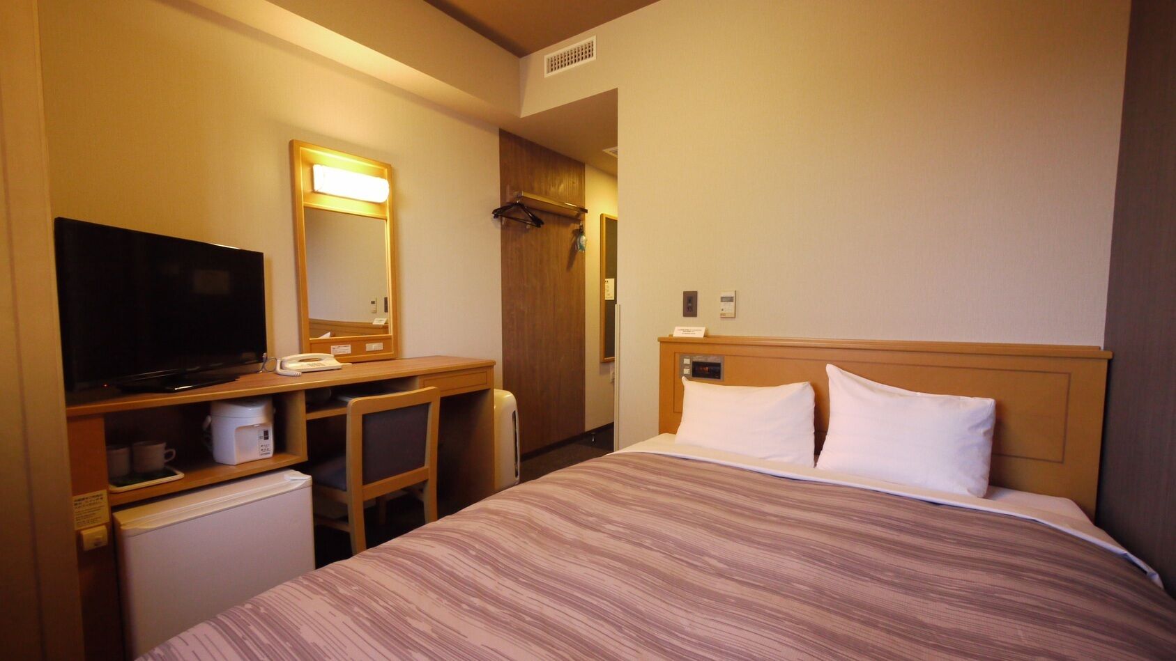 A hotel room with a bed, a desk with a television, a mirror, a wardrobe, and a small refrigerator.