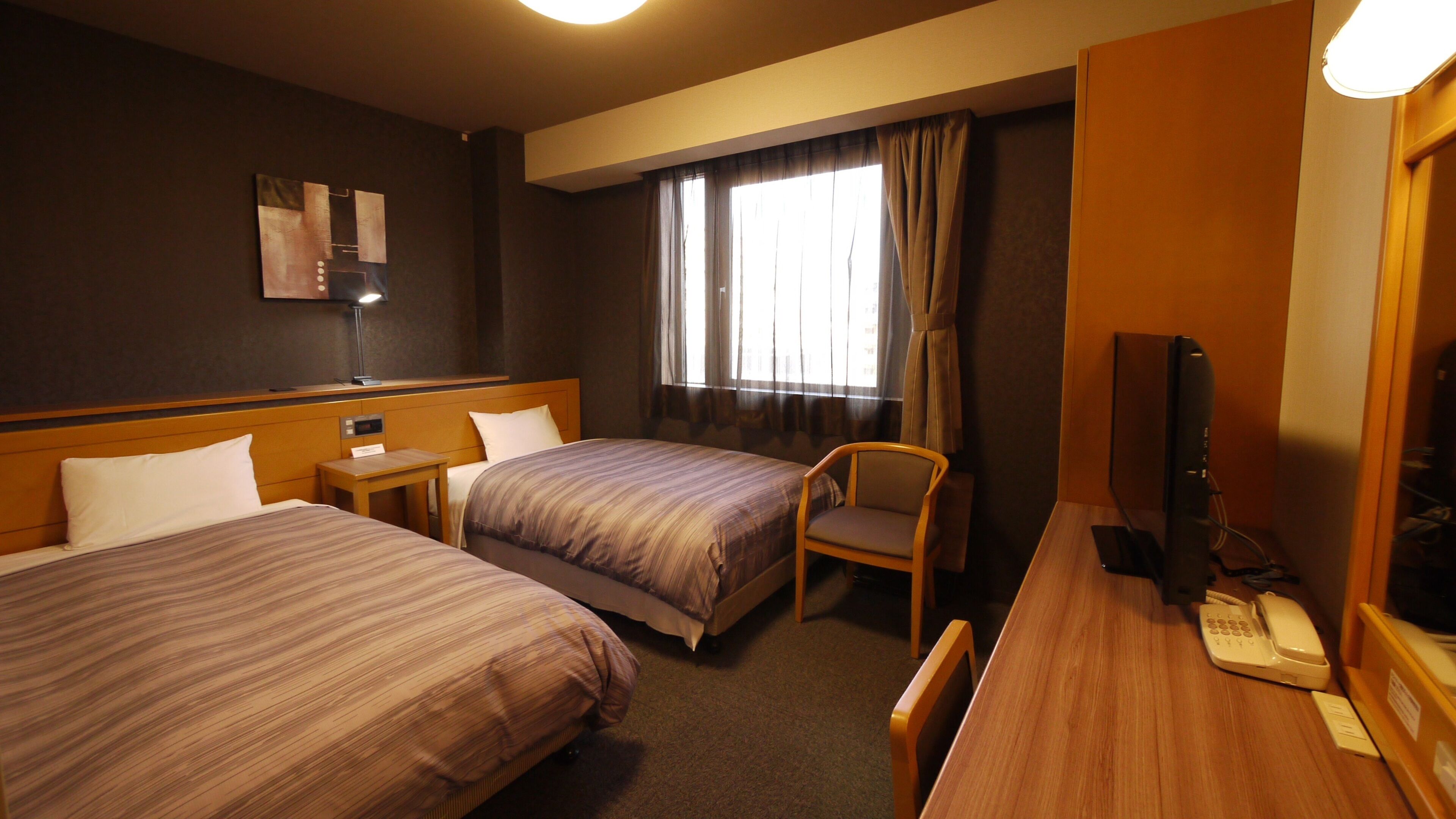 Quarto de hotel com duas camas, uma escrivaninha, telefone, cadeira, televisão e janela com cortinas.