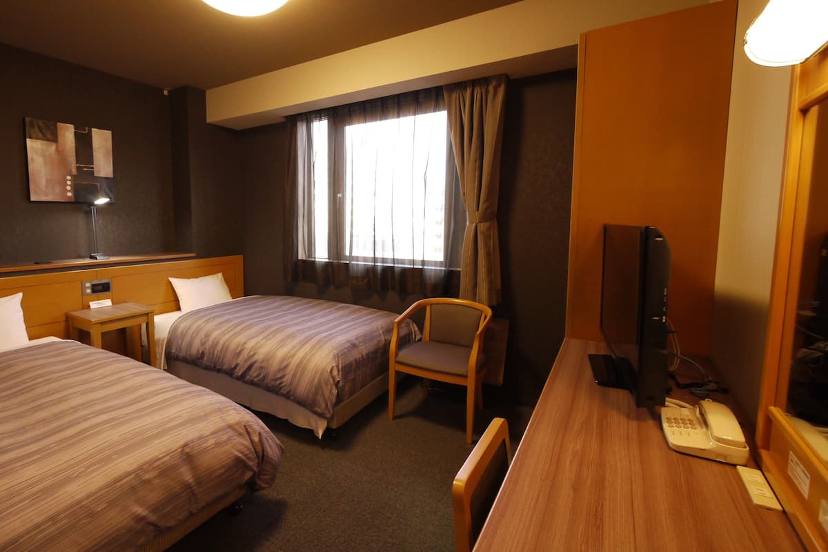 Quarto de hotel com duas camas, uma escrivaninha, telefone, cadeira, televisão e janela com cortinas.