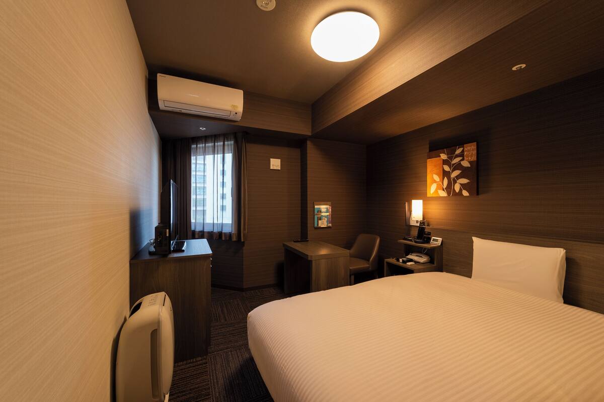 A hotel room with a bed, desk, chair, and a small table with a lamp.