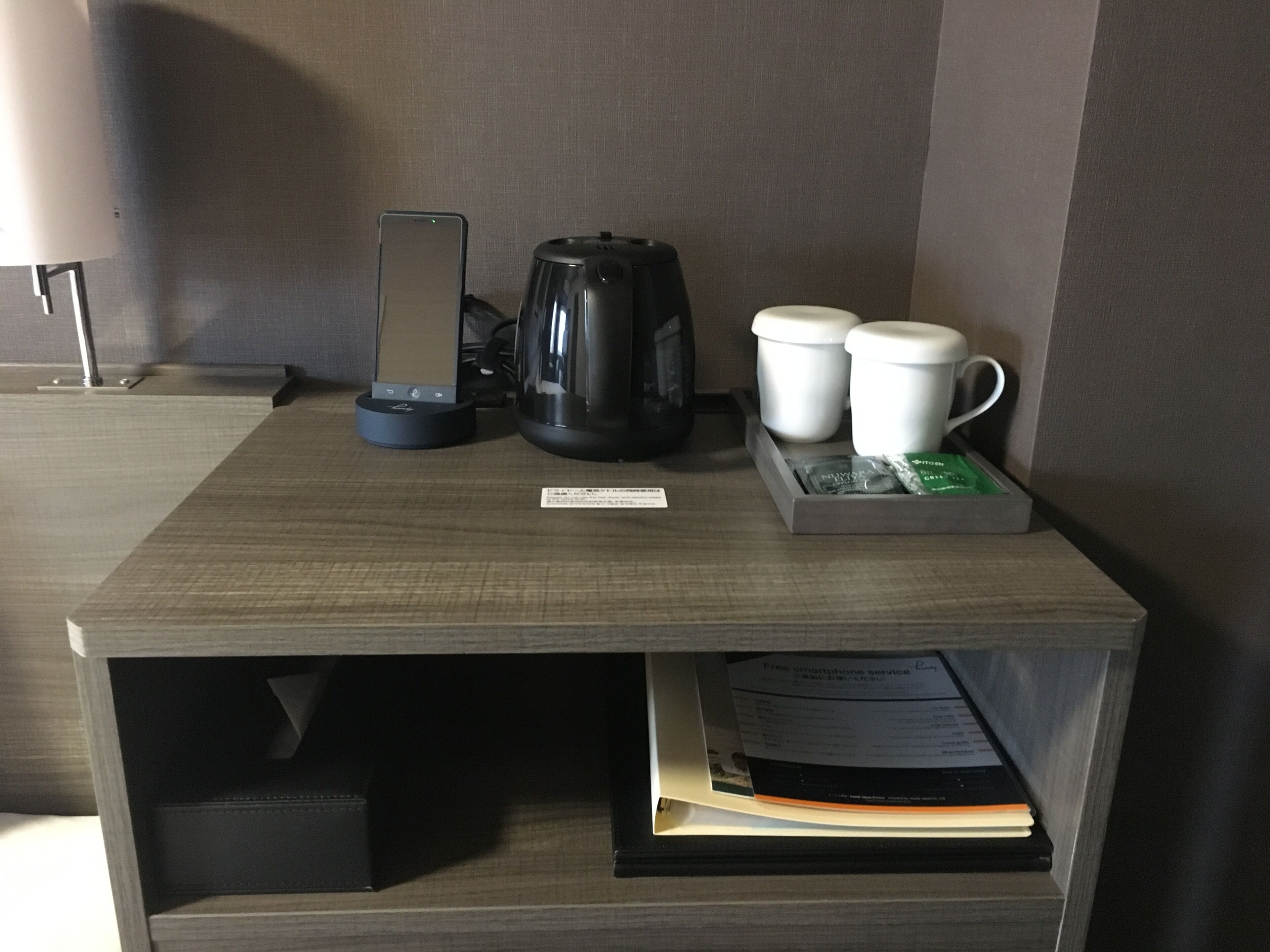 A hotel room with a wooden nightstand, a smartphone charging station, a coffee maker, two white cups, and a tray with a packet of tissues.