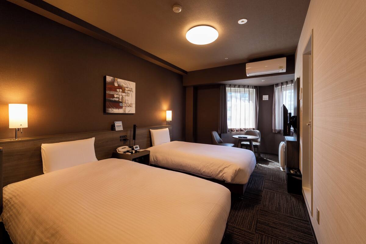 A hotel room with two beds, a desk with a chair, a small table, a lamp, and a painting on the wall.