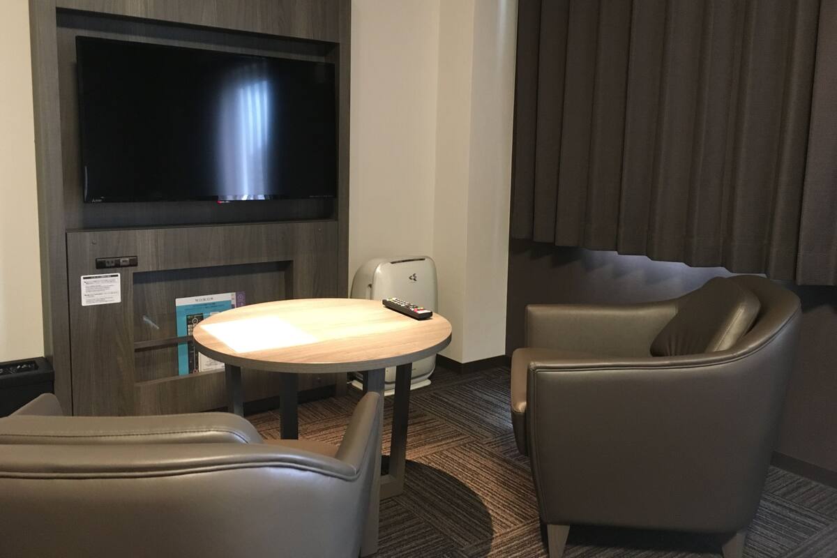 A hotel room with a flat-screen TV mounted on the wall, a small round table with a remote control, two leather chairs, and a dark curtain.
