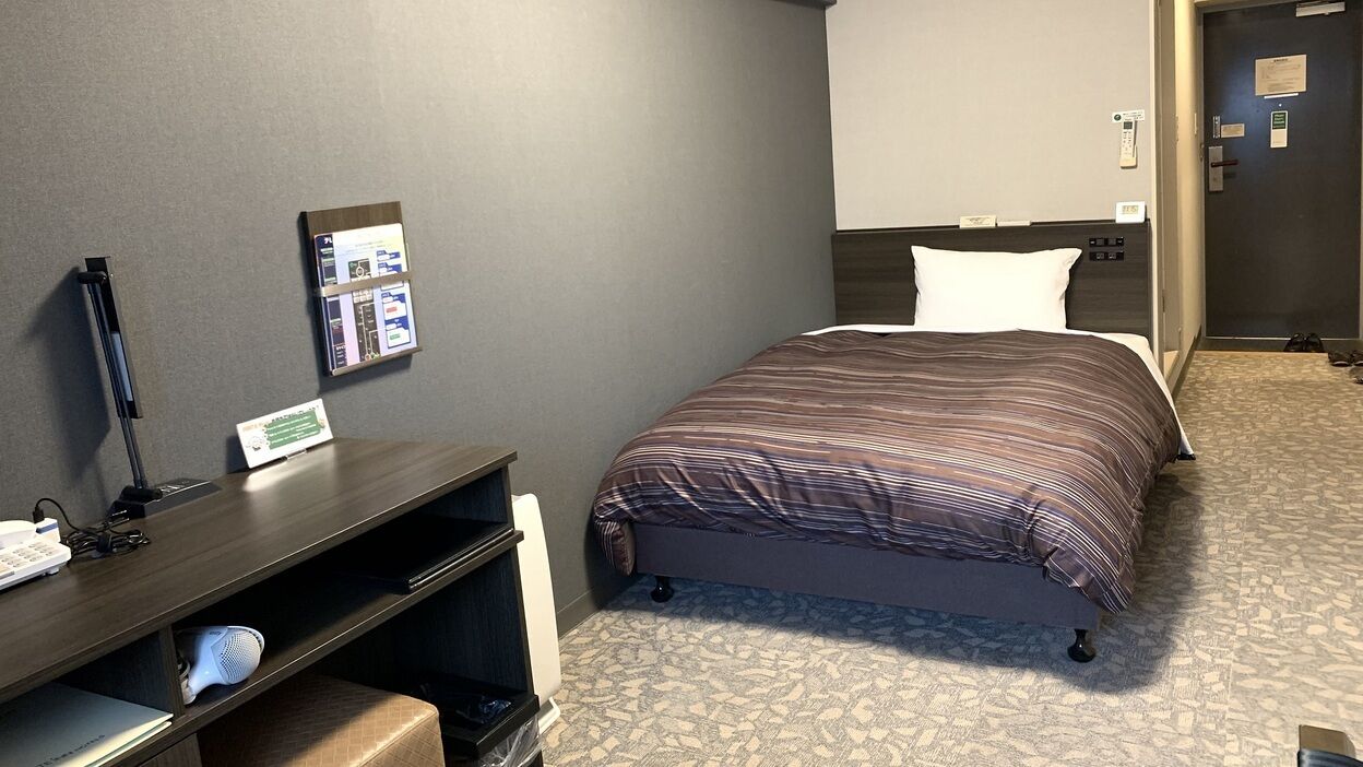 A hotel room with a bed, a desk with a computer, a telephone, and a bulletin board.