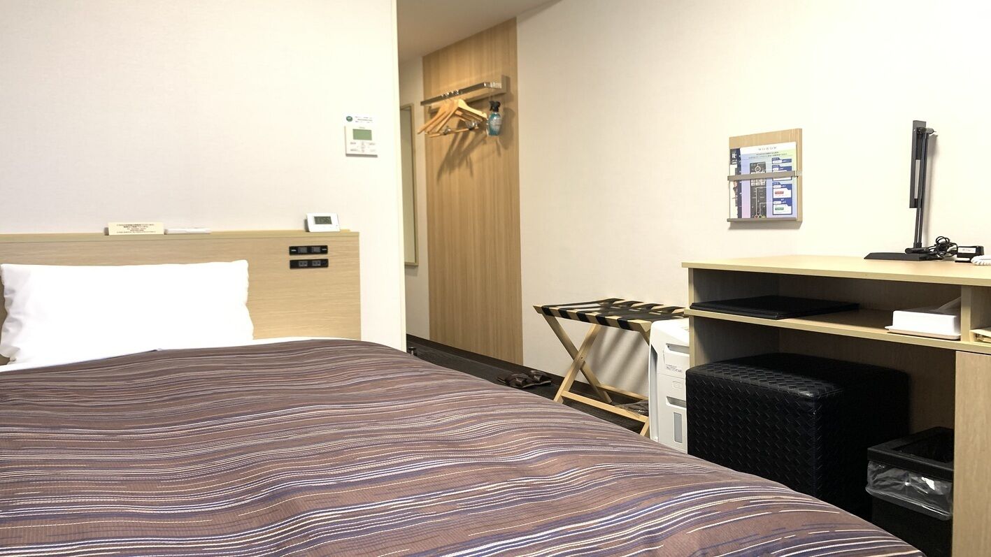 A hotel room with a bed, a desk with a computer, a chair, and a trash bin.