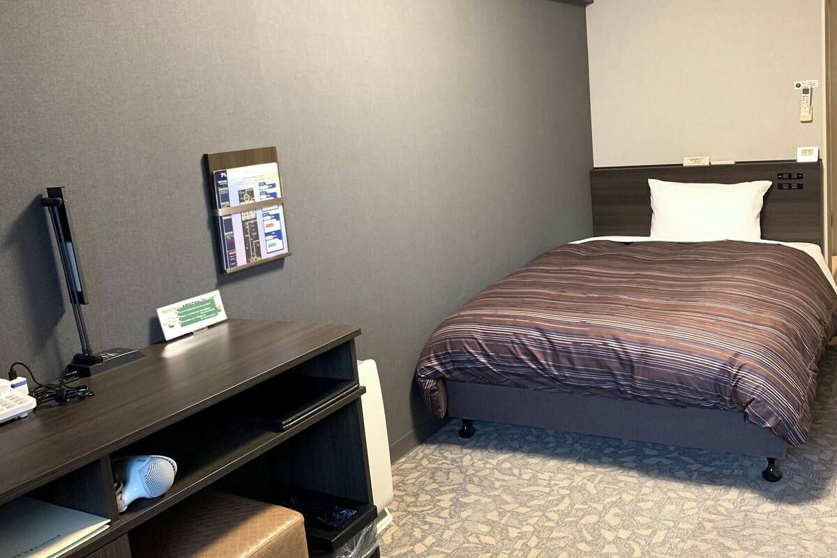 Ein Hotelzimmer mit Bett, Schreibtisch mit Computer, Telefon und einer an der Wand befestigten Infotafel.