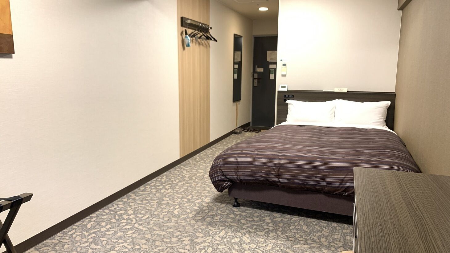 A hotel room with a bed, a desk, a coat rack, and a door.