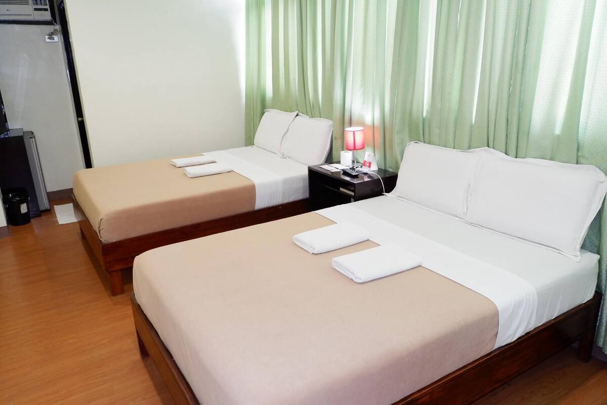A hotel room with two beds, each with a beige bedspread and white pillows. There is a nightstand with a lamp and a phone, and a small trash bin to the left.