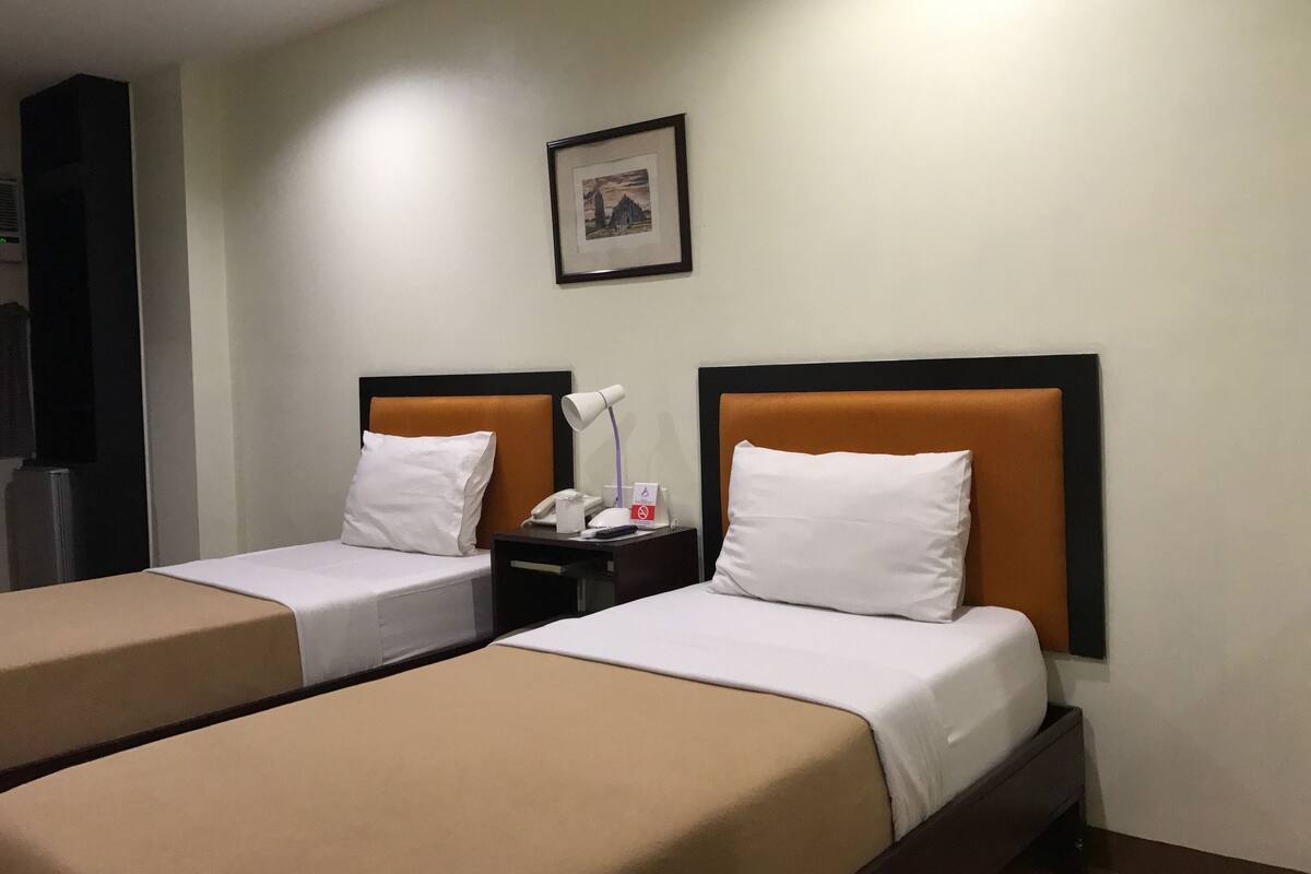 A hotel room with two beds, each with a white pillow and a beige bedspread. There is a nightstand with a lamp and a phone between the beds. A framed picture hangs on the wall above the beds.